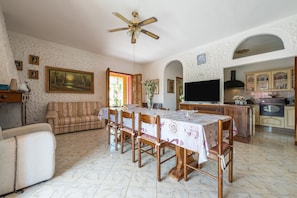A dining area with a table set for six, a sofa, a television, and a kitchen in the background. - Villa 'Chiara' with Mountain View, Wi-Fi and Air Conditioning (Quartu Sant'Elena)