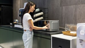 A woman in a white shirt and jeans operating a microwave in a modern kitchen with grey tiles and a black countertop. - Hotel Daily (Ansan)