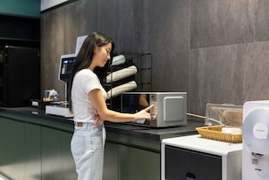 Una mujer con camisa blanca y jeans usando un microondas en una cocina moderna con azulejos grises y una encimera negra.