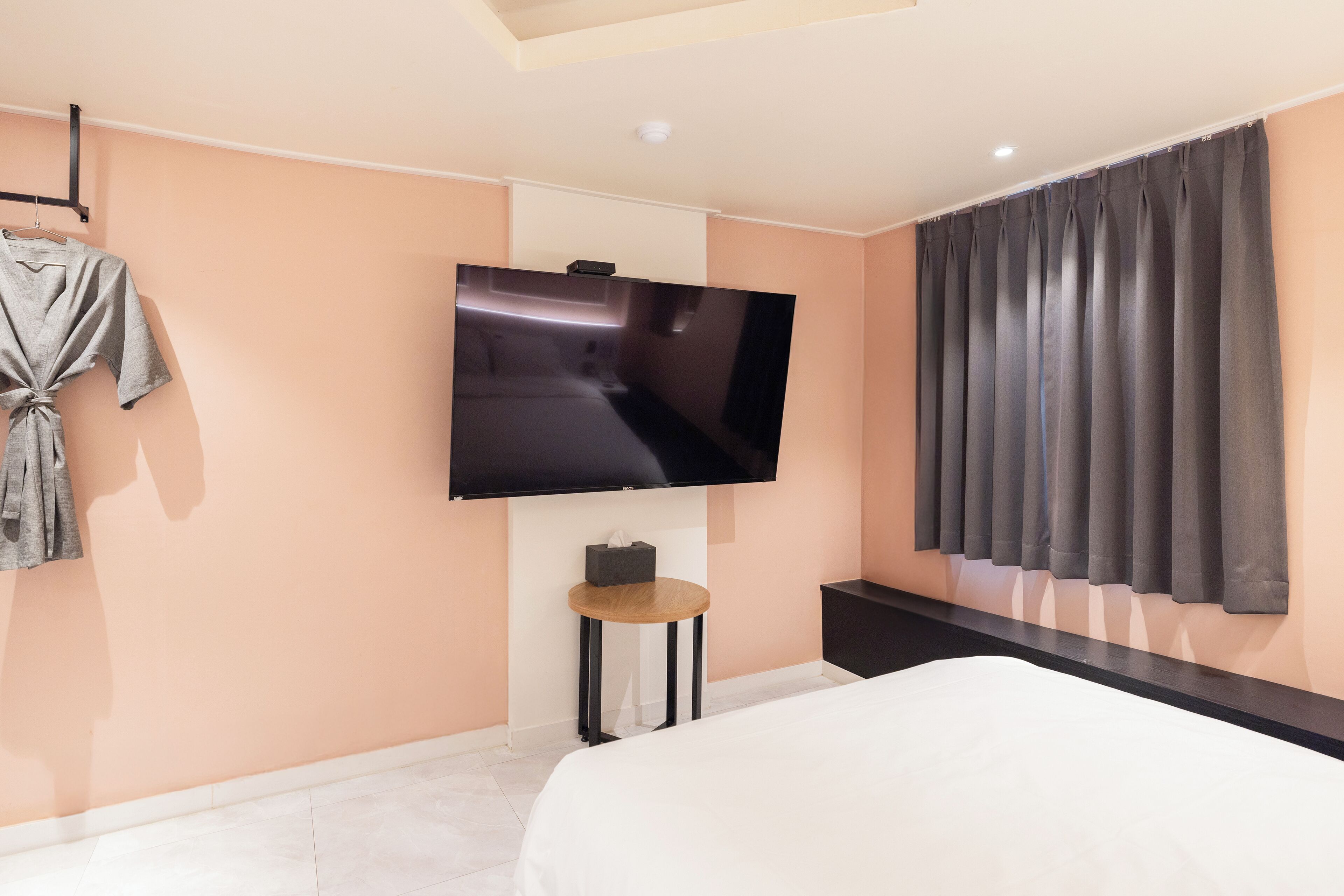 A modern hotel room with a large flat-screen TV mounted on the wall, a bed with white bedding, a small round table with a tissue box, and a dark-colored bench.