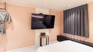 A modern hotel room with a large flat-screen TV mounted on the wall, a bed with white bedding, a small round table with a tissue box, and a dark-colored bench.
