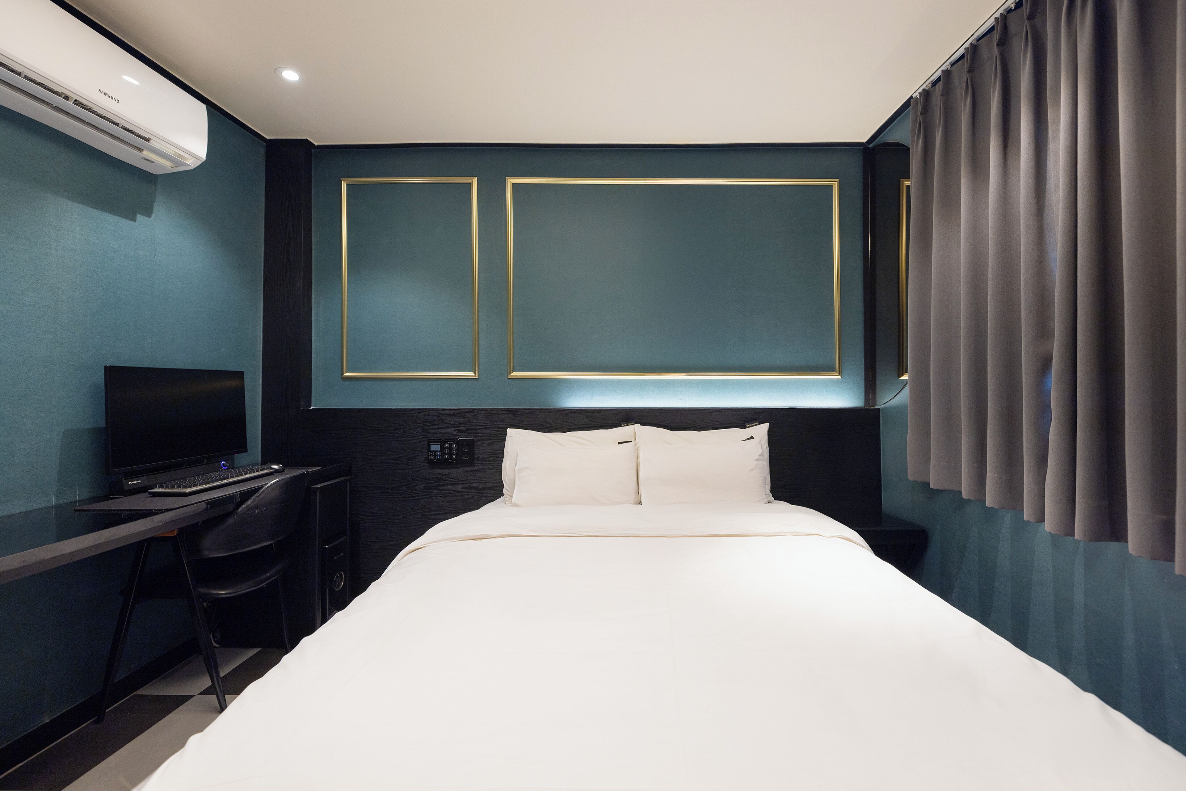 A hotel room with a bed, a desk with a computer, and an air conditioning unit.