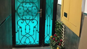 A hallway with a black door featuring intricate designs and a frosted glass panel. A yellow door is visible to the right, and a potted plant with flowers is placed near the wall. - Studio with terrace 10 mn from airport and beach sablette beatch (El-Harrach)
