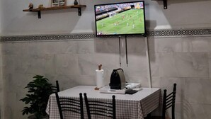 A dining area with a table set for four, a television mounted on the wall displaying a sports event, and decorative shelves above. - Studio with terrace 10 mn from airport and beach sablette beatch (El-Harrach)