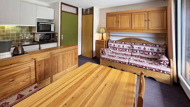 A compact kitchen with a microwave, stove, and a green door. There is a wooden dining table with chairs and a sofa with patterned cushions.