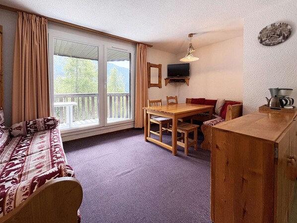 A cozy room with a sofa, a dining table, chairs, a television, and a kitchen area with a kettle.