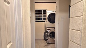 Un cuarto de lavandería con lavadoras y secadoras apiladas, una ventana y una puerta que da a otra habitación.