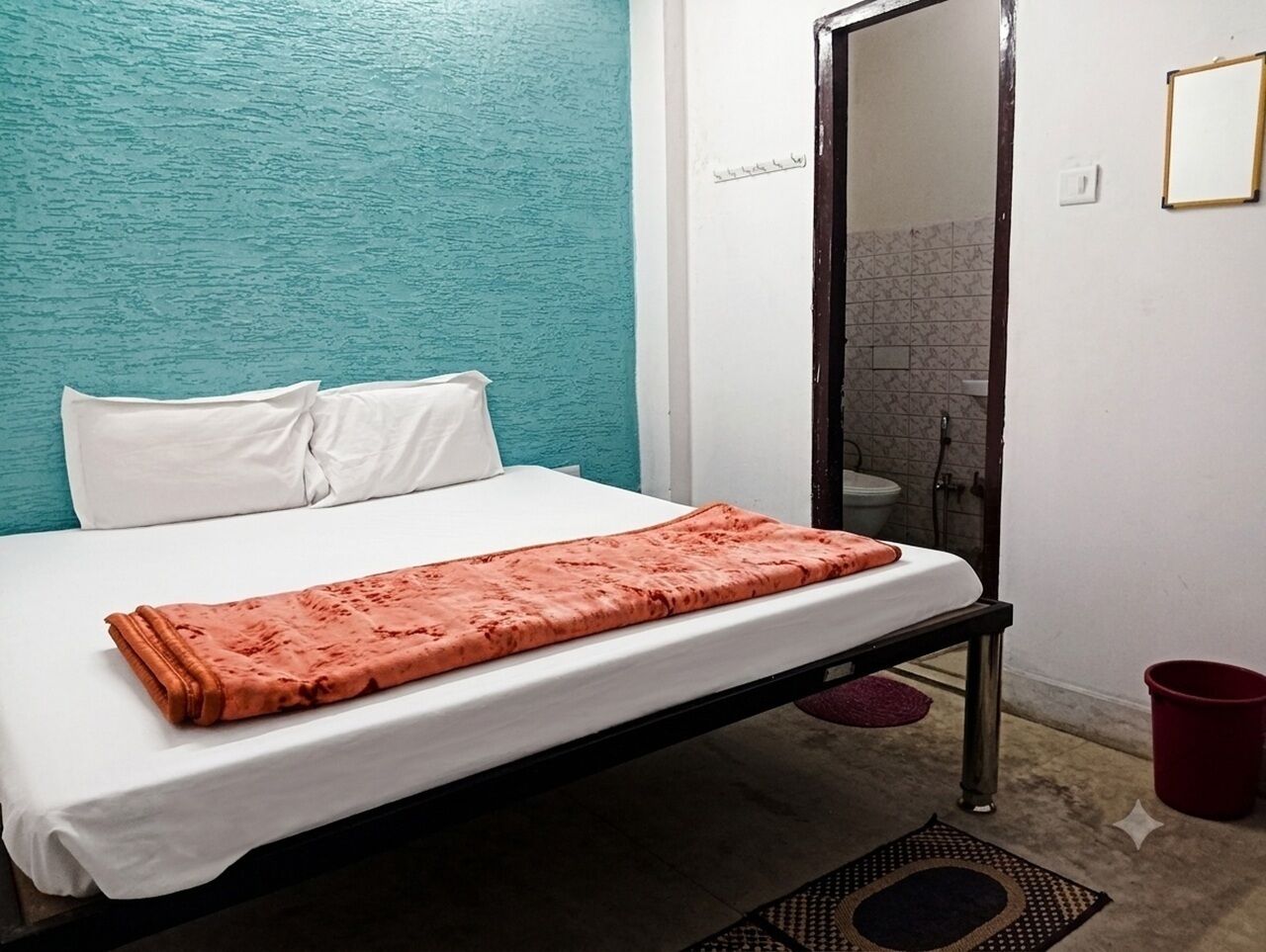 A single bed with a white mattress and a pink blanket, a small red bucket, and a mirror on the wall.