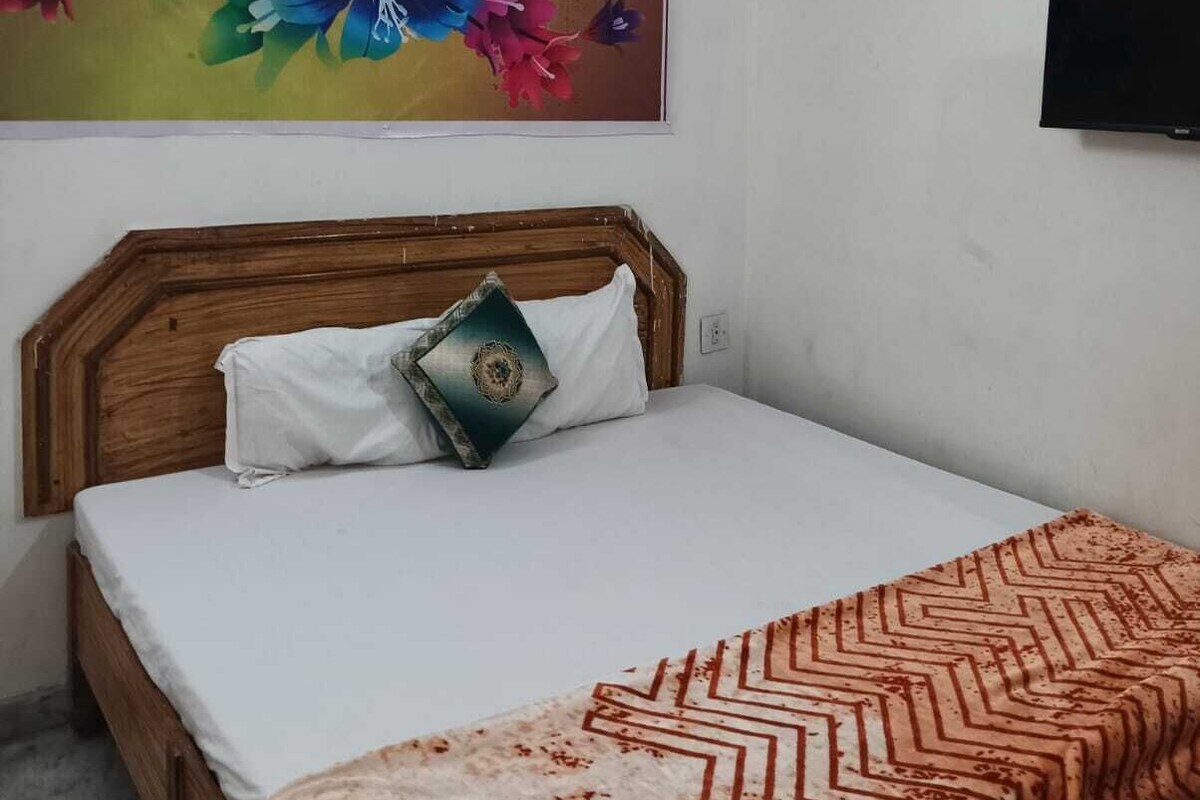 Een slaapkamer met een houten hoofdbord, een bed met wit beddengoed, een kleurrijk bloemenkunstwerk aan de muur, en een aan de muur bevestigde televisie.