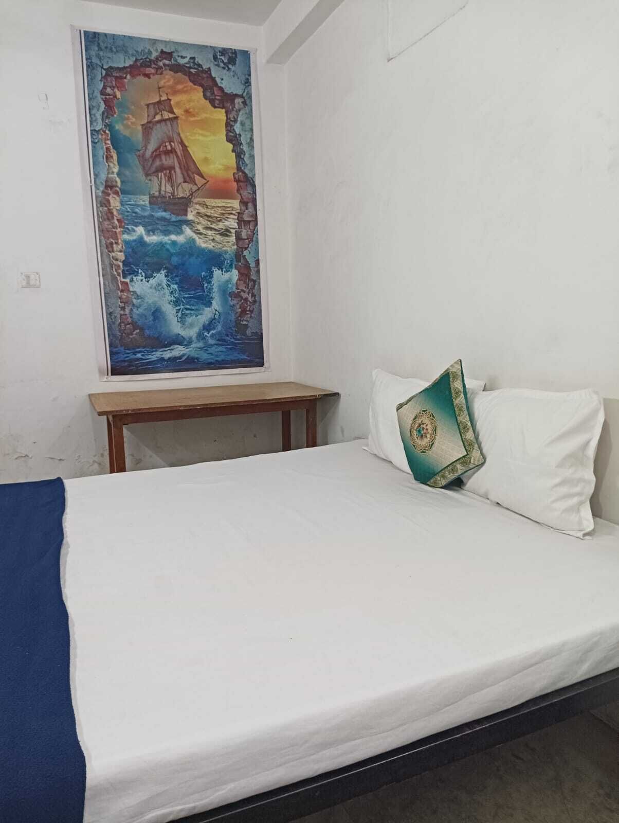 Una habitación con una cama, un cuadro de un barco en un acantilado y una pequeña mesa de madera.