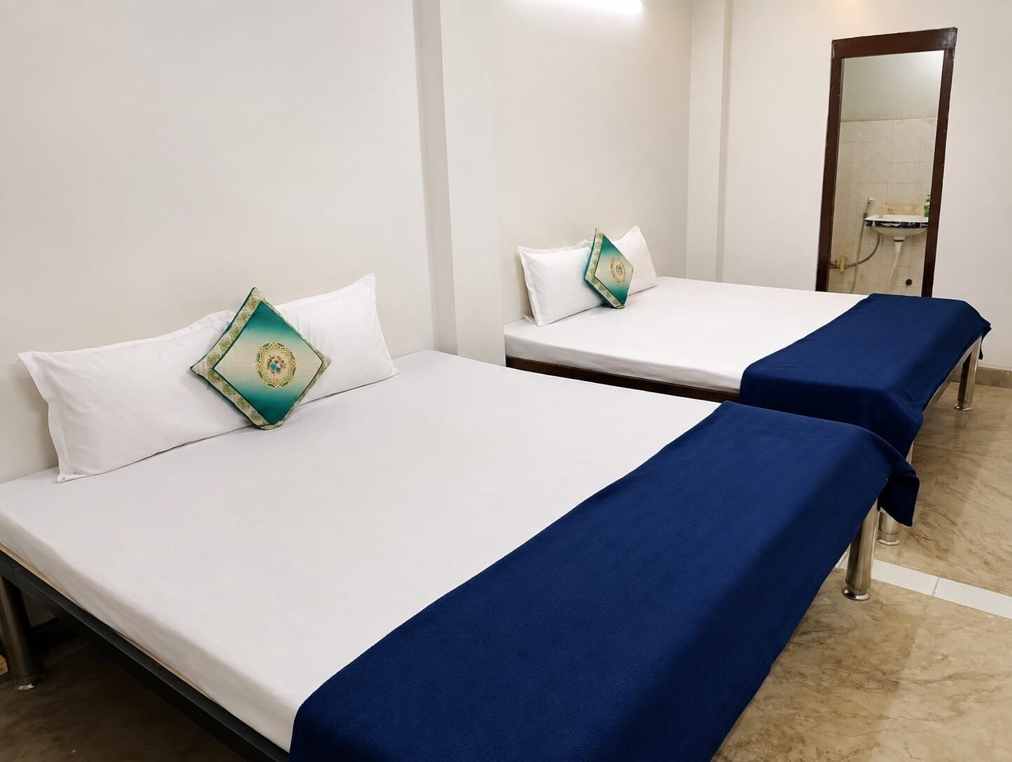 Deux lits simples avec du linge de lit blanc et des couvre-lits bleus, un mur blanc et une salle de bain avec un lavabo et un miroir.