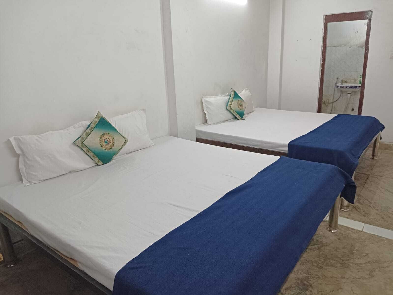 Two single beds with white linens and blue bedspreads in a room with white walls and a tiled floor.