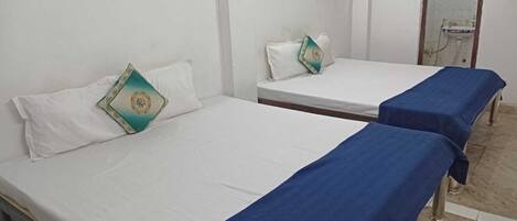 Two single beds with white linens and blue bedspreads in a room with white walls and a tiled floor.