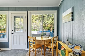 Une pièce avec une table et des chaises en bois, une porte et une vue sur une clôture bleue et des arbres à l’extérieur.