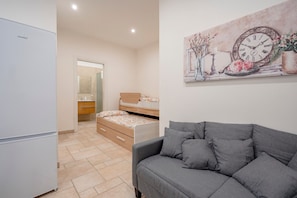 A modern bedroom with a grey sofa, a bed with a floral duvet, a painting of a clock, and a white refrigerator.