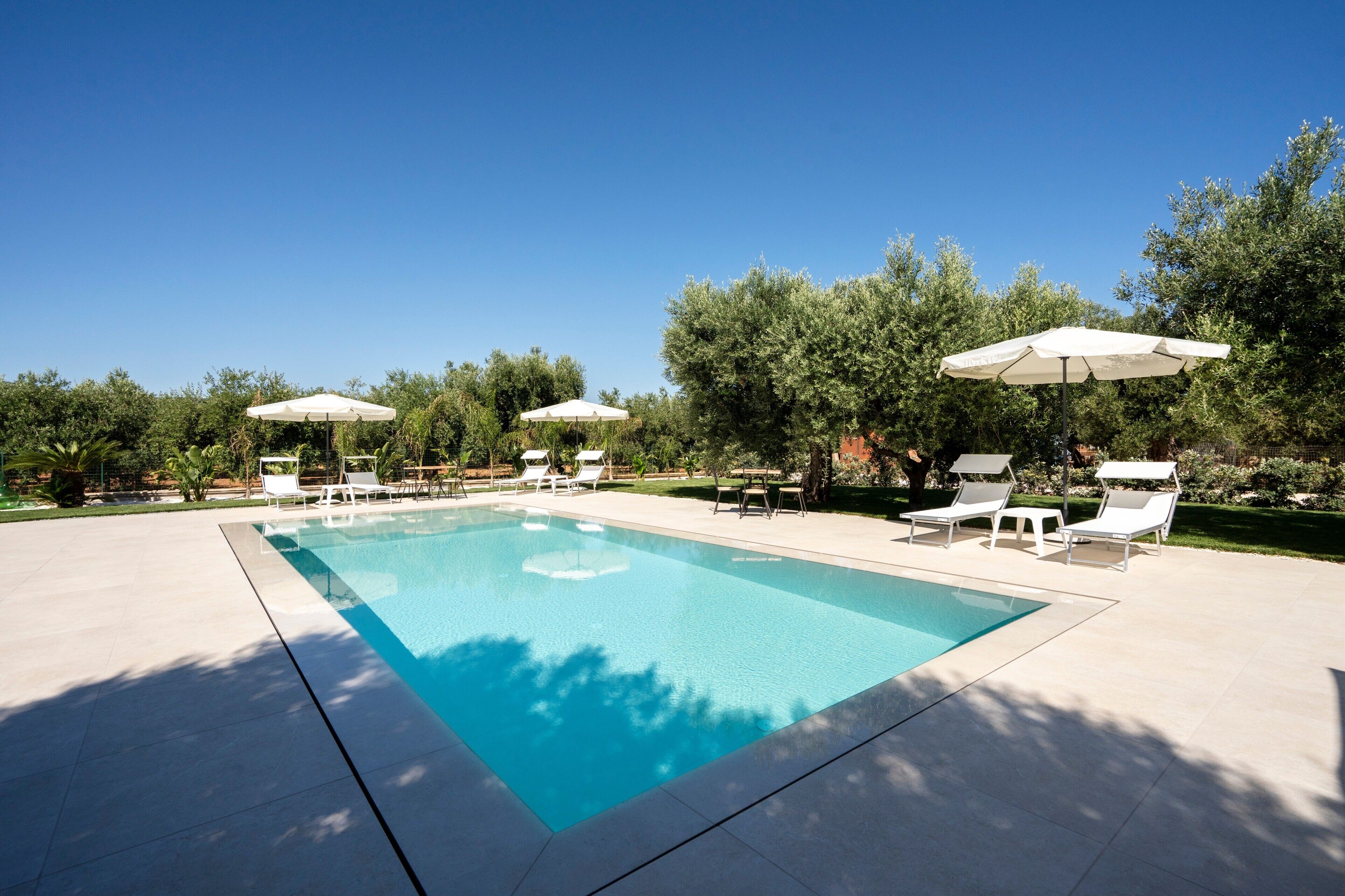 Una piscina rectangular con sillas de descanso blancas y sombrillas, rodeada de árboles y un cielo despejado.