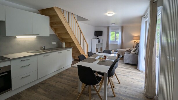 Eine moderne Küche mit weißen Schränken, einer Holztreppe, einem Essbereich mit Tisch und Stühlen sowie einem Wohnbereich mit einer Couch und Vorhängen.