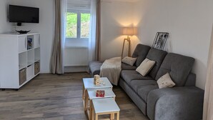 Ein Wohnzimmer mit grauem Sofa, einem weißen TV-Schrank, einem kleinen Tisch und einer Stehlampe.