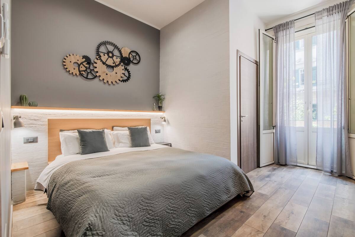 Un dormitorio moderno con una cama grande, cabecera, un reloj fijado a la pared y una puerta corrediza de vidrio al exterior.