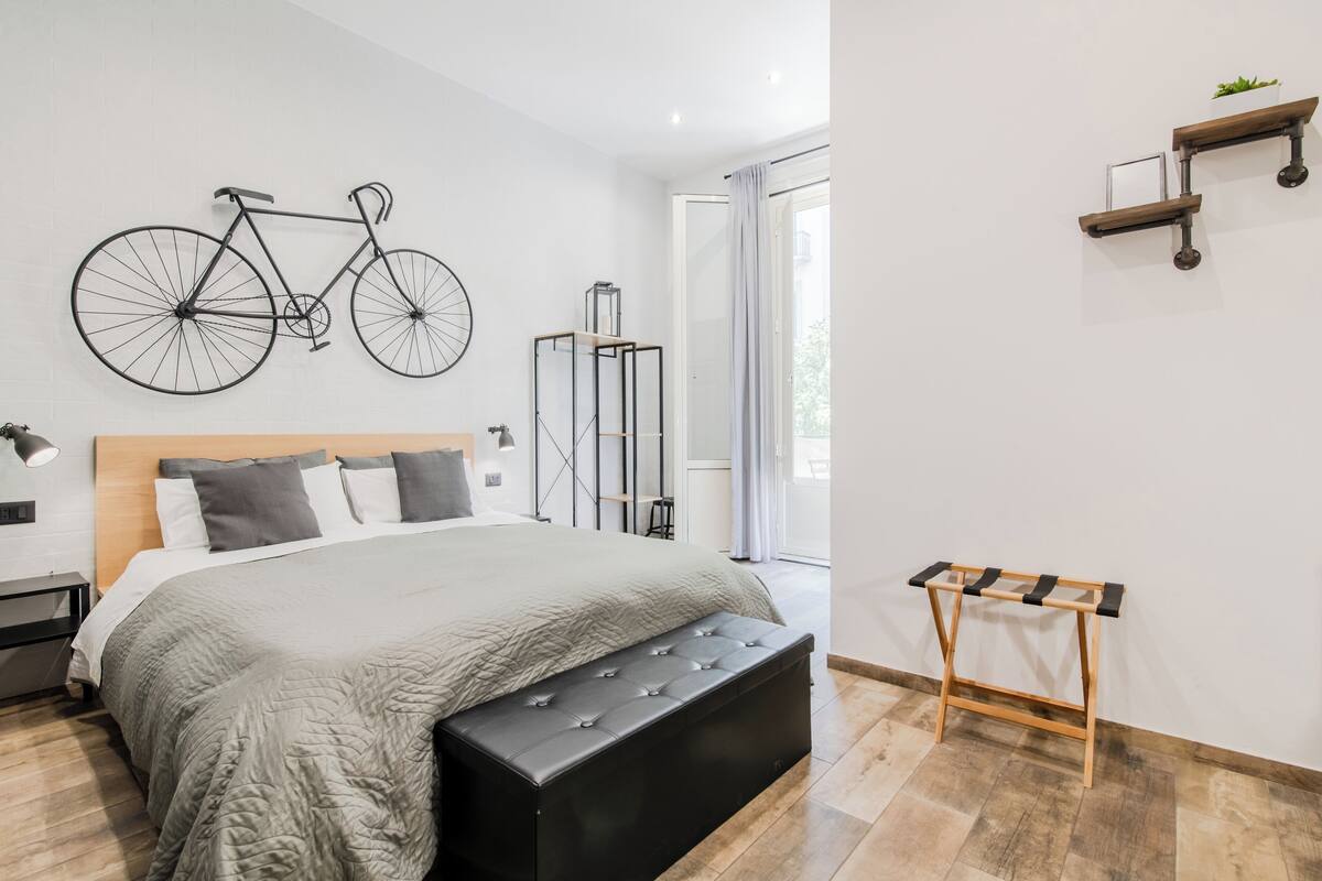Un dormitorio moderno con una cama grande, un banco de madera, un cuadro de bicicleta en la pared, una lámpara de pie y una ventana con cortinas.