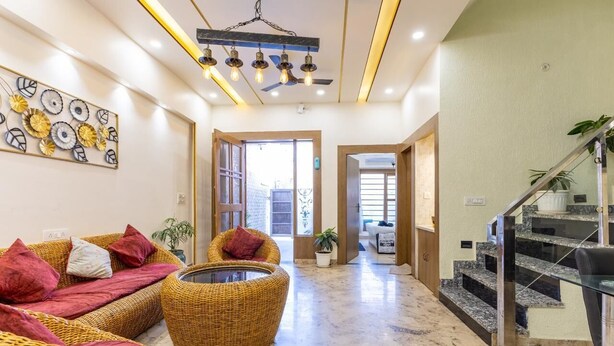 A spacious living area with a sofa set, a coffee table, and a wicker chair. There is a staircase with a glass railing leading to an upper floor. - A radiant villa on the Ganges (Rishikesh)