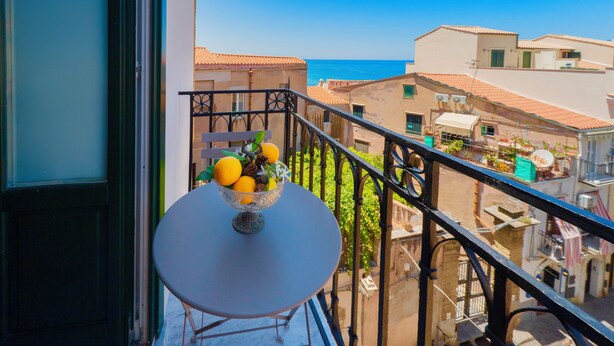 A balcony with a table and a bowl of fruit, overlooking a coastal town with terracotta-roofed buildings.