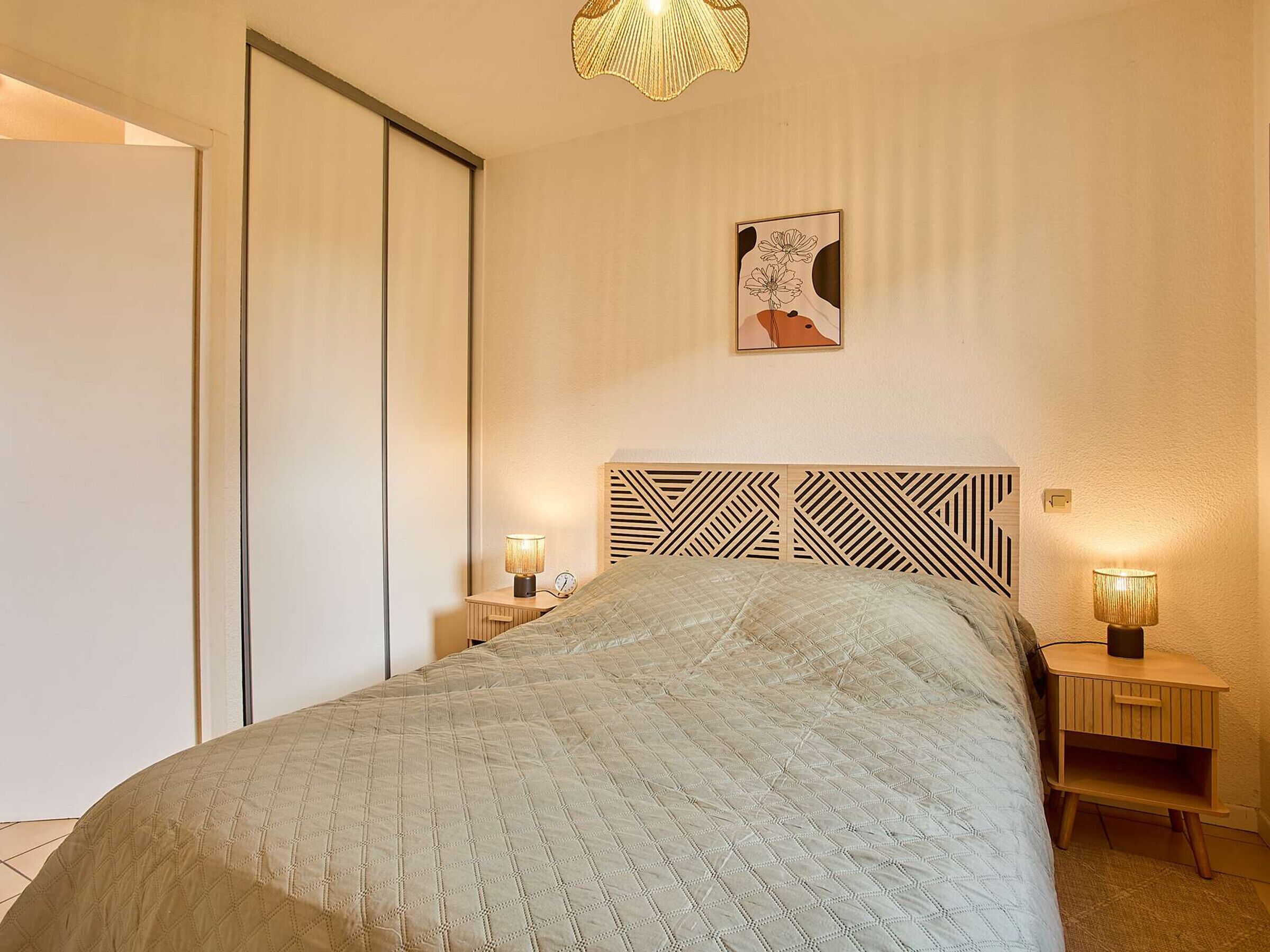 A bedroom with a bed, two bedside tables with lamps, a wall-mounted artwork, and a ceiling light fixture.