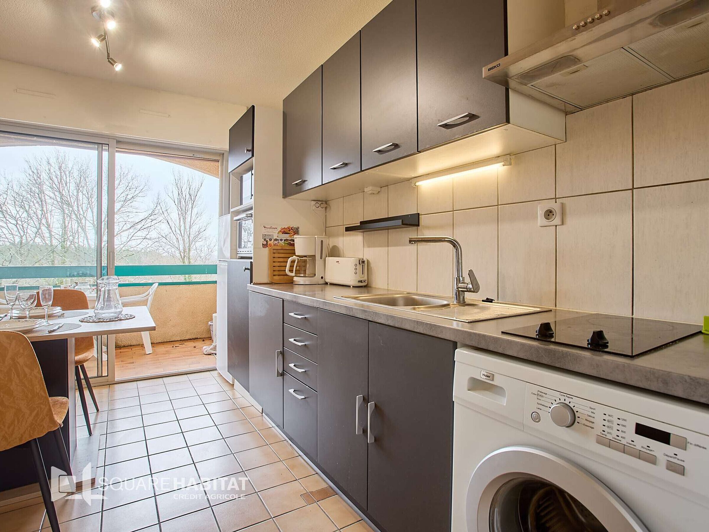 A modern kitchen with grey cabinets, a built-in washing machine, and a dining area with a table and chairs.