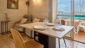 A dining area with a table set for four, a sofa, and a view through a glass door to the outside. - Calm T2 Apartment with Balcony and Wifi (Barbotan-les-Thermes)