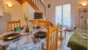 Un comedor con una mesa dispuesta para cuatro, un televisor sobre un soporte, una escalera y vistas a través de un ventanal.