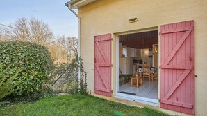 Una casa con un pequeño patio, una cocina visible a través de un vidrio y un arbusto grande al frente.