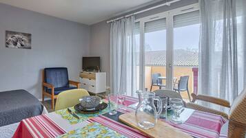 Une pièce confortable avec un lit, une chaise, une table recouverte d’une nappe à rayures et un téléviseur posé sur un meuble blanc.