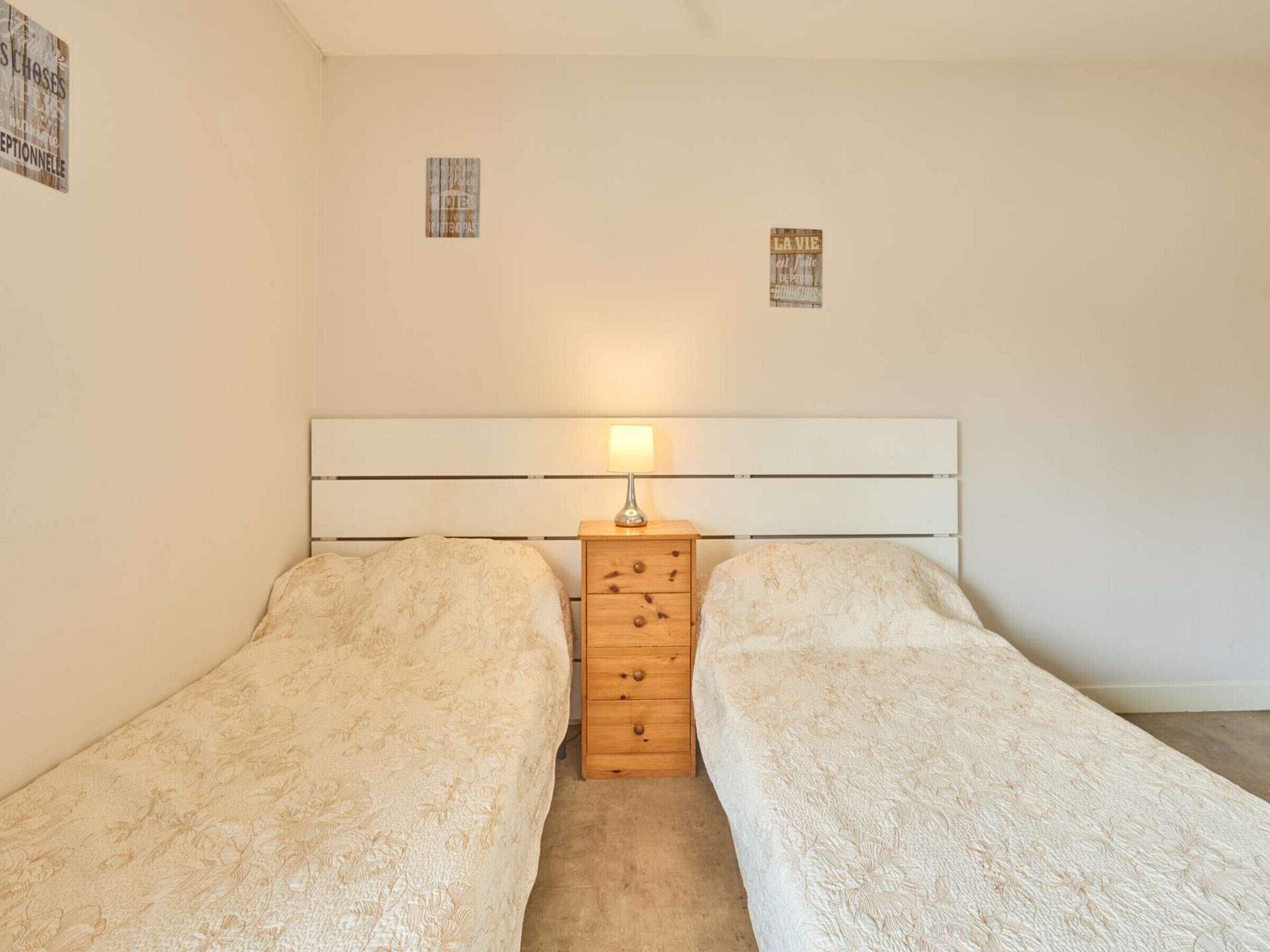 Un dormitorio con dos camas individuales, una mesita de noche de madera con cajones, una lámpara y dos pequeños carteles en la pared.