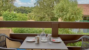 Une table de balcon, dressée avec des assiettes, une carafe et des verres, d’où l’on peut admirer un paysage verdoyant et luxuriant ainsi qu’un bâtiment doté d’un toit en tuiles.