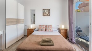 A bedroom with a bed, a nightstand, a bedside table, a window with a view, and a sliding door to a balcony.