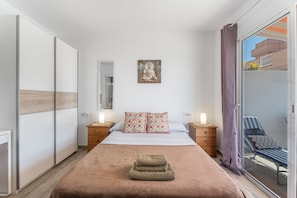 A bedroom with a bed, a nightstand, a bedside table, a window with a view, and a sliding door to a balcony.