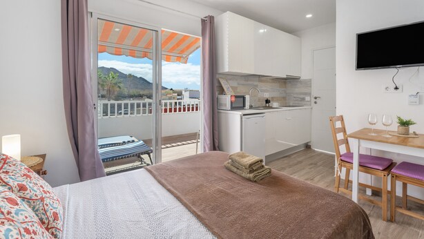 A bedroom with a bed, a desk with a chair, a TV, and a view of a balcony with a kitchen.