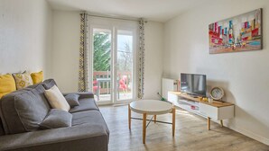 A living room with a grey sofa, a round table, a TV on a wooden stand, and a colorful painting on the wall. - Spacious renovated T2 with balcony, wifi and pets allowed in Barbotan-les-Thermes (Barbotan-les-Thermes)