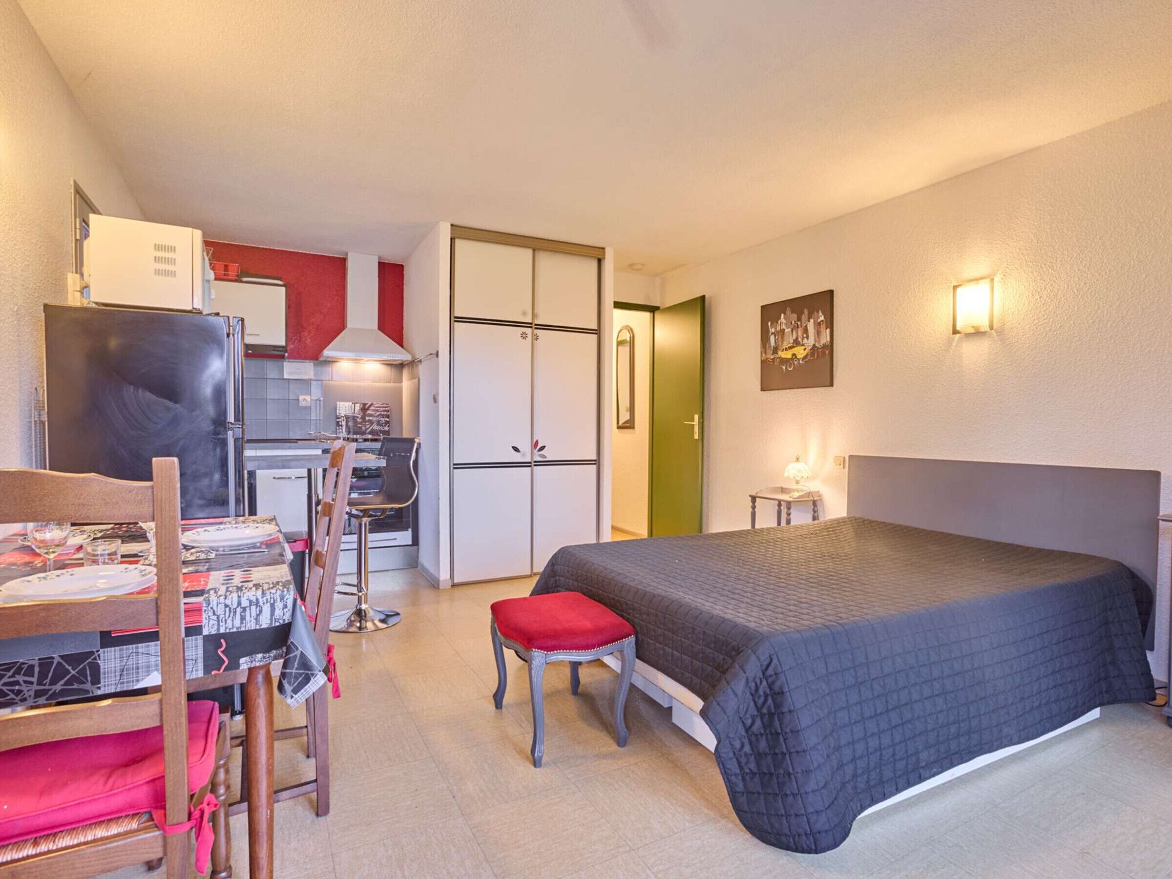 Quarto de hotel compacto com cama, escrivaninha com cadeira, cozinha compacta e armário.