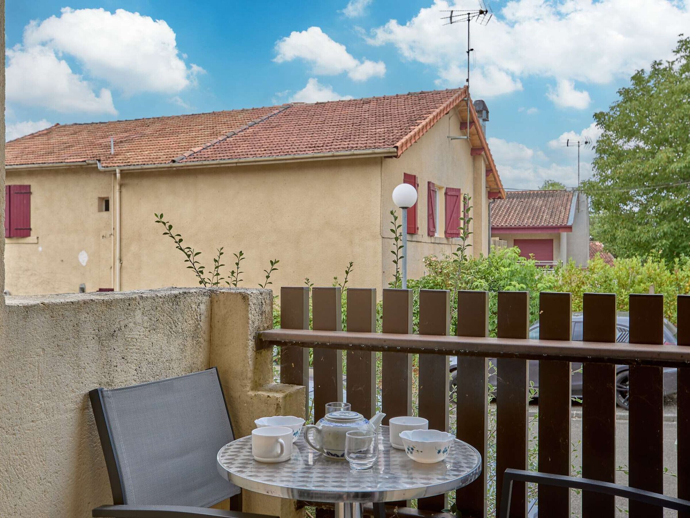 A balcony with a table set for tea, a chair, and a view of a building with red shutters and a tiled roof.