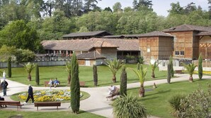 Un bâtiment traditionnel en bois, doté d’un toit de chaume, entouré d’un jardin bien entretenu avec des bancs et des allées piétonnes.