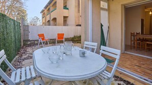 An outdoor patio with a round table and white chairs, a brick entrance, and a building with balconies in the background. - Charming T1 bis with private garden in Barbotan-les-Thermes (Barbotan-les-Thermes)