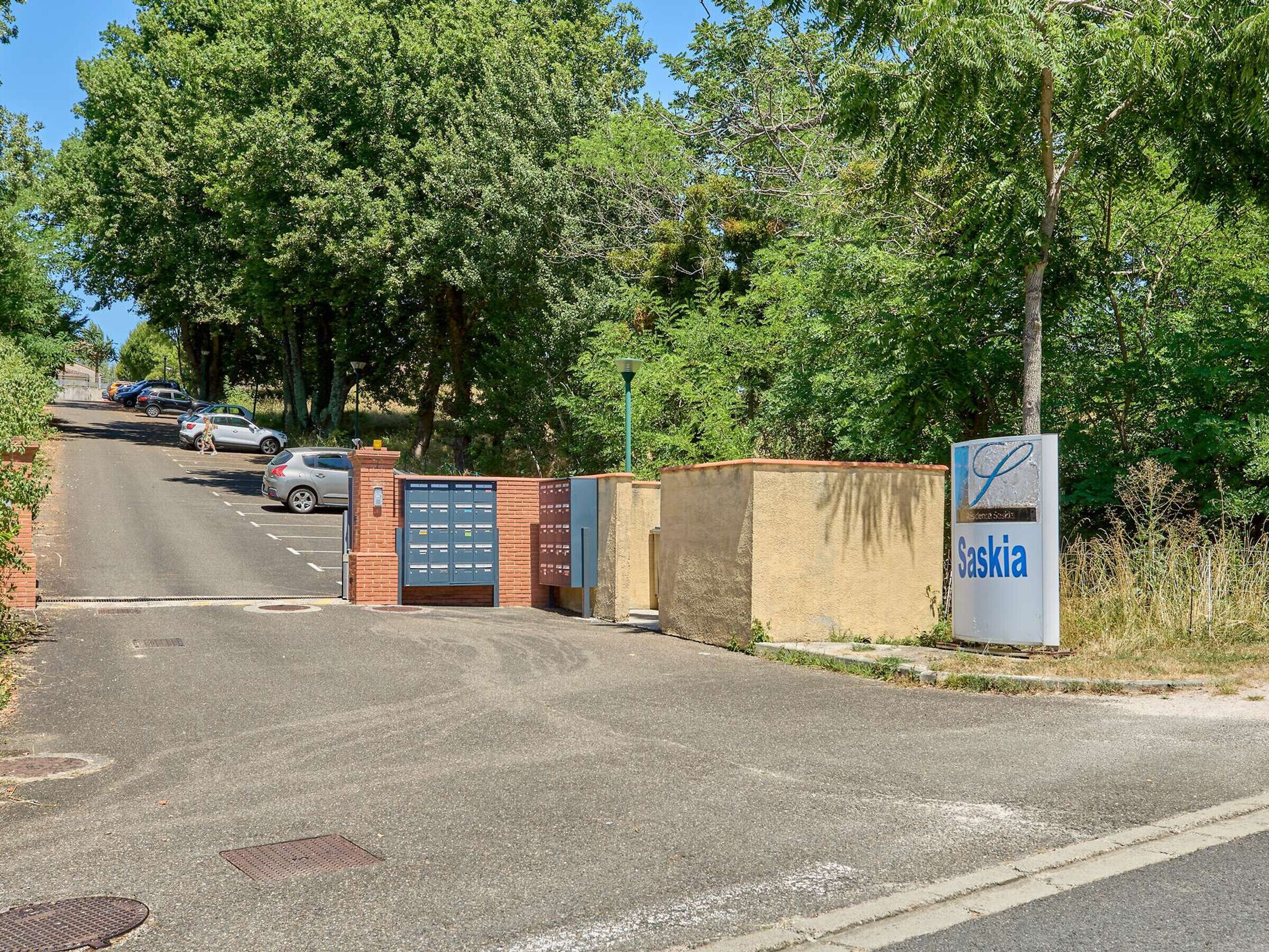 Un espace de stationnement doté d’une entrée fermée par une barrière, d’un panneau indiquant « Saskia », et d’une route qui monte.