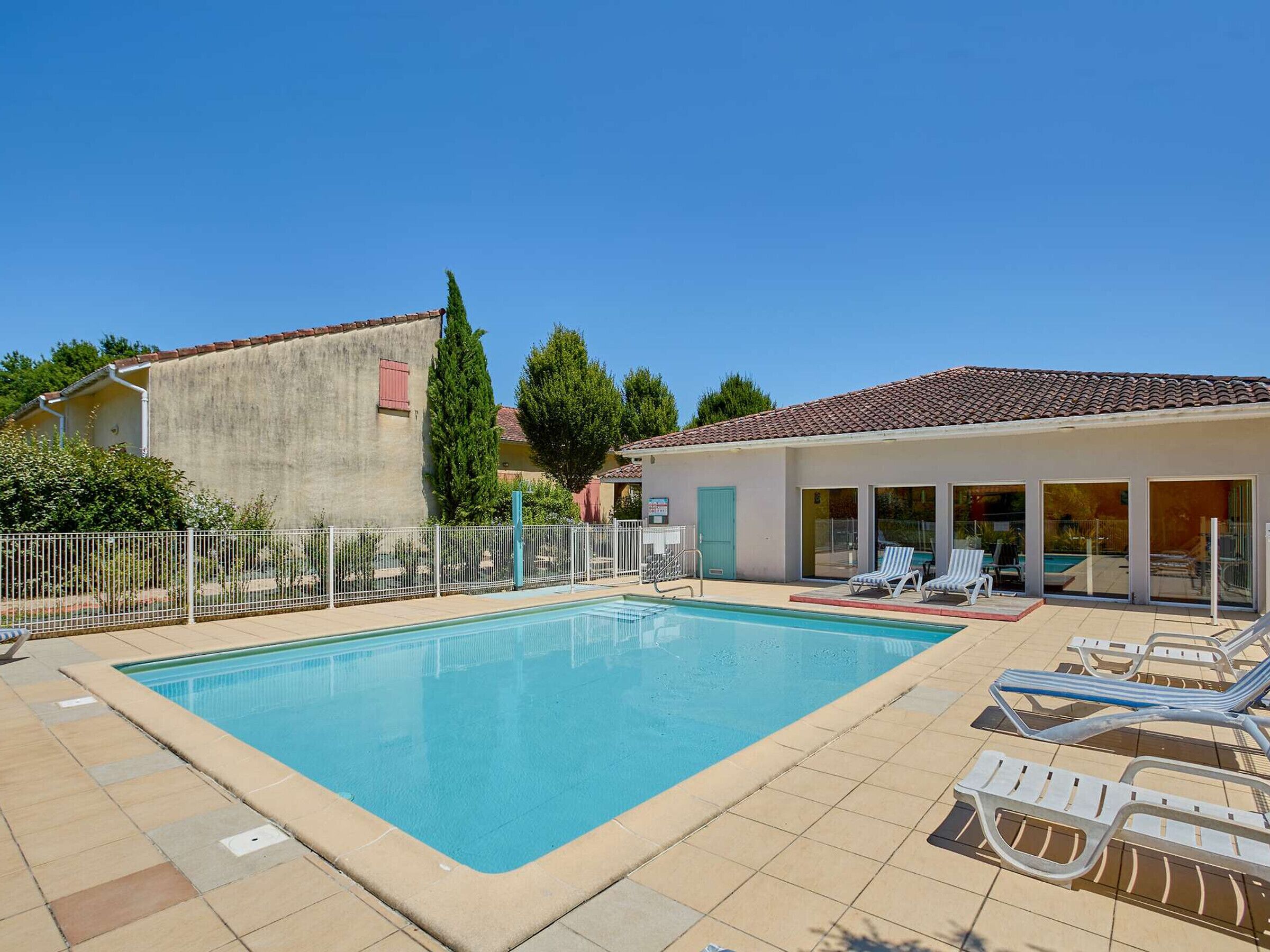A rectangular swimming pool with clear blue water, surrounded by a tiled deck. There are several lounge chairs and a covered area with glass doors.
