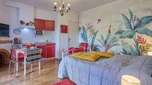 A bedroom with a bed, a red chair, a red sofa, a kitchen area, and a mural of tropical plants.