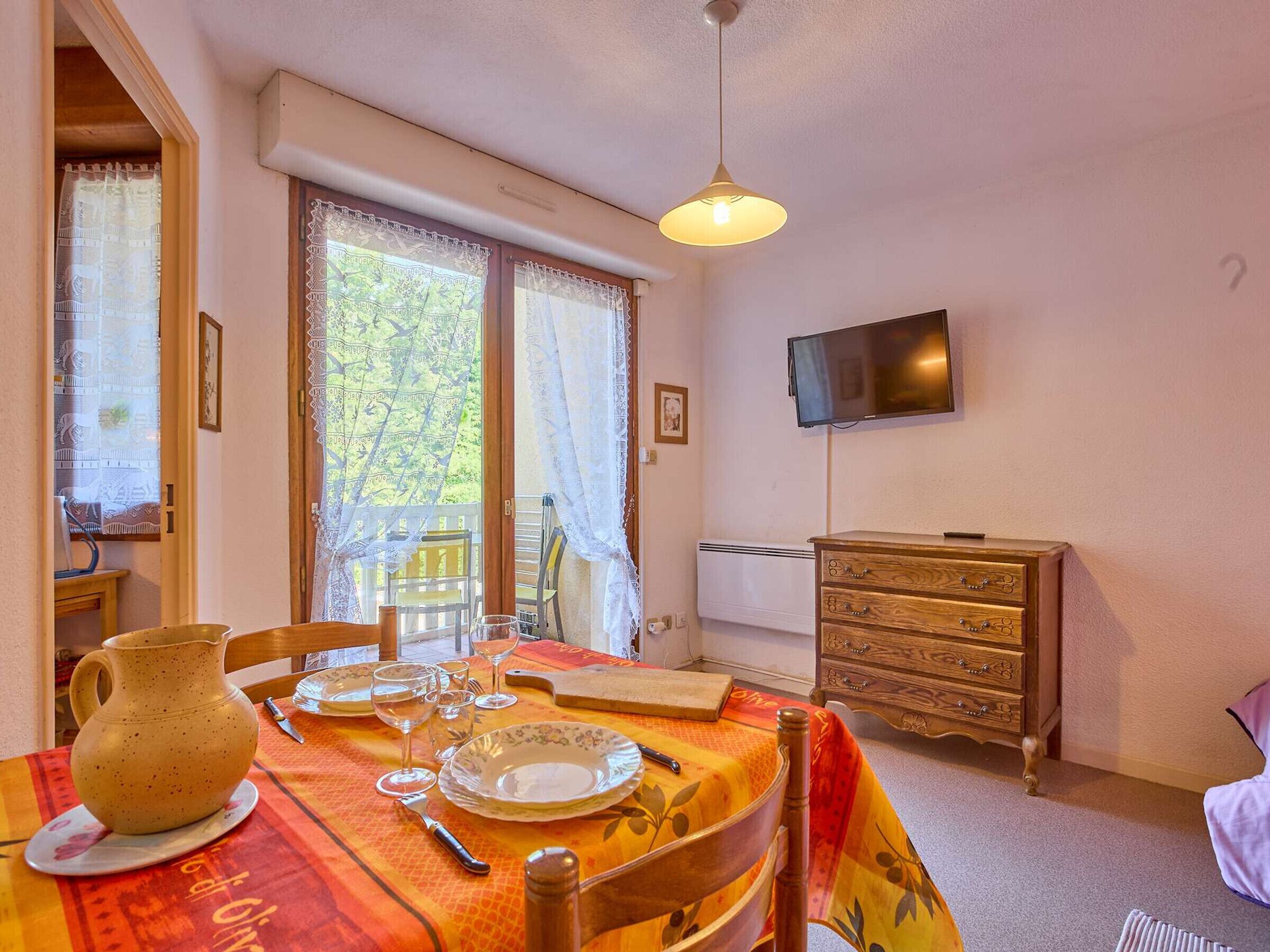 A dining area with a table set for two, a wooden cabinet, and a television on the wall.