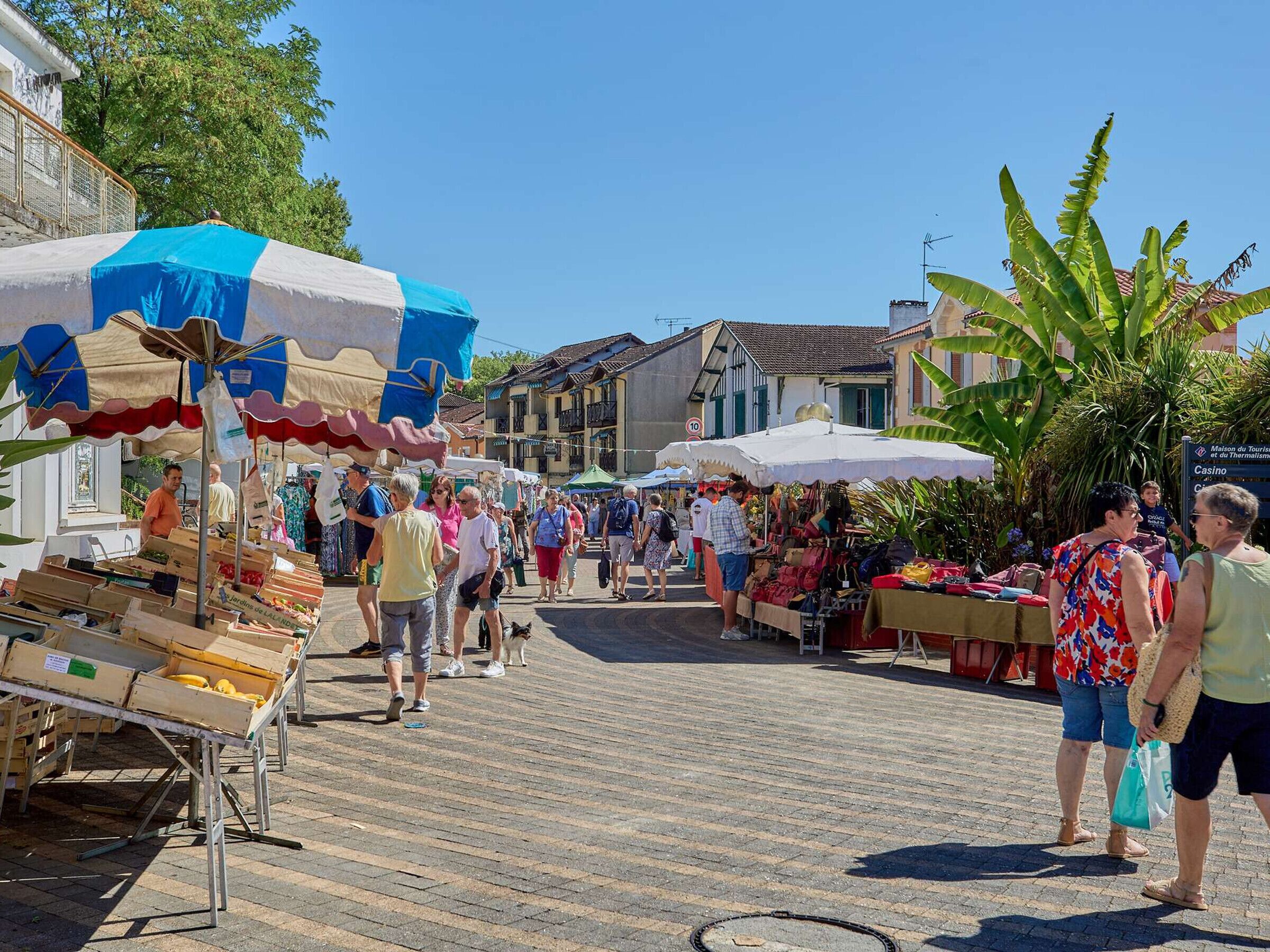A bustling outdoor market with vendors and shoppers, wooden stalls, and a clear blue sky.