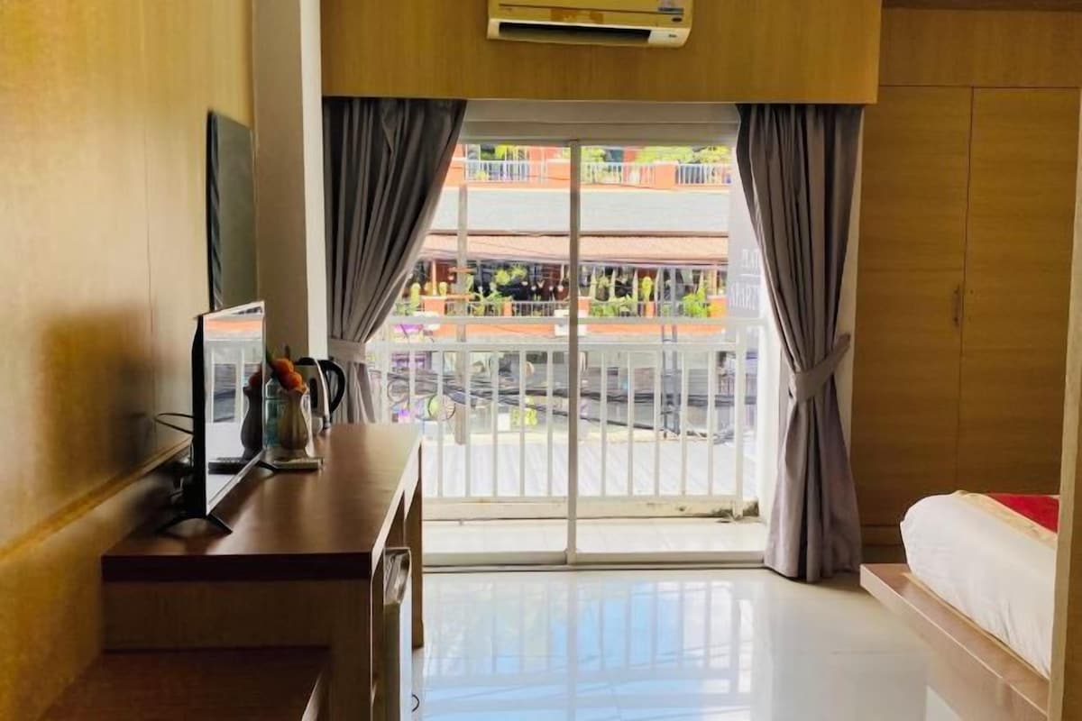 A hotel room with a bed, a desk with a chair, a window with curtains, and a view of a balcony with outdoor furniture.