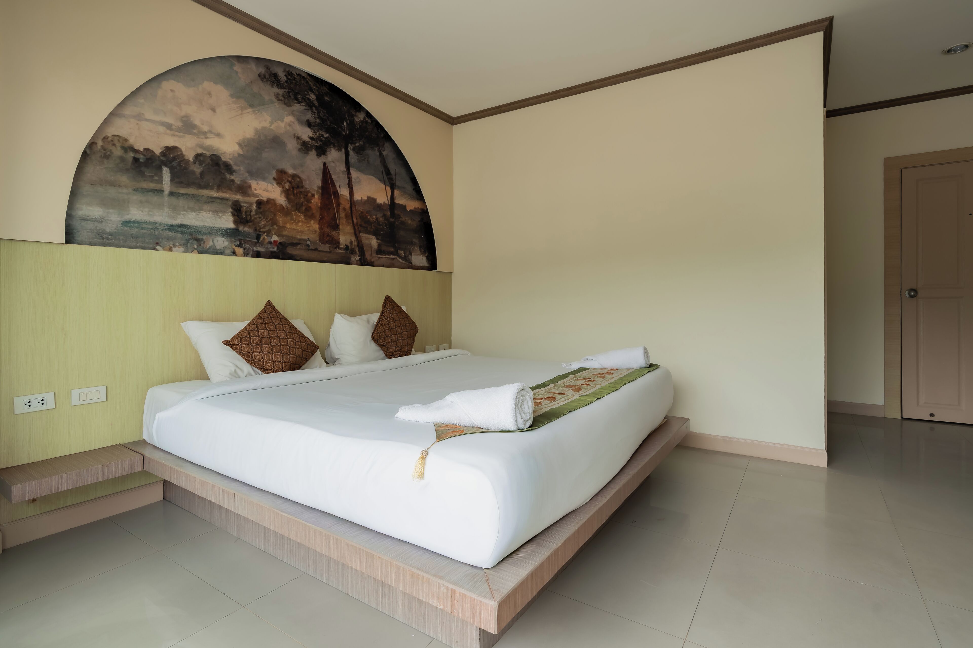 A neatly made bed with a white comforter and pillows, a large landscape painting on the wall, and a door to the right.