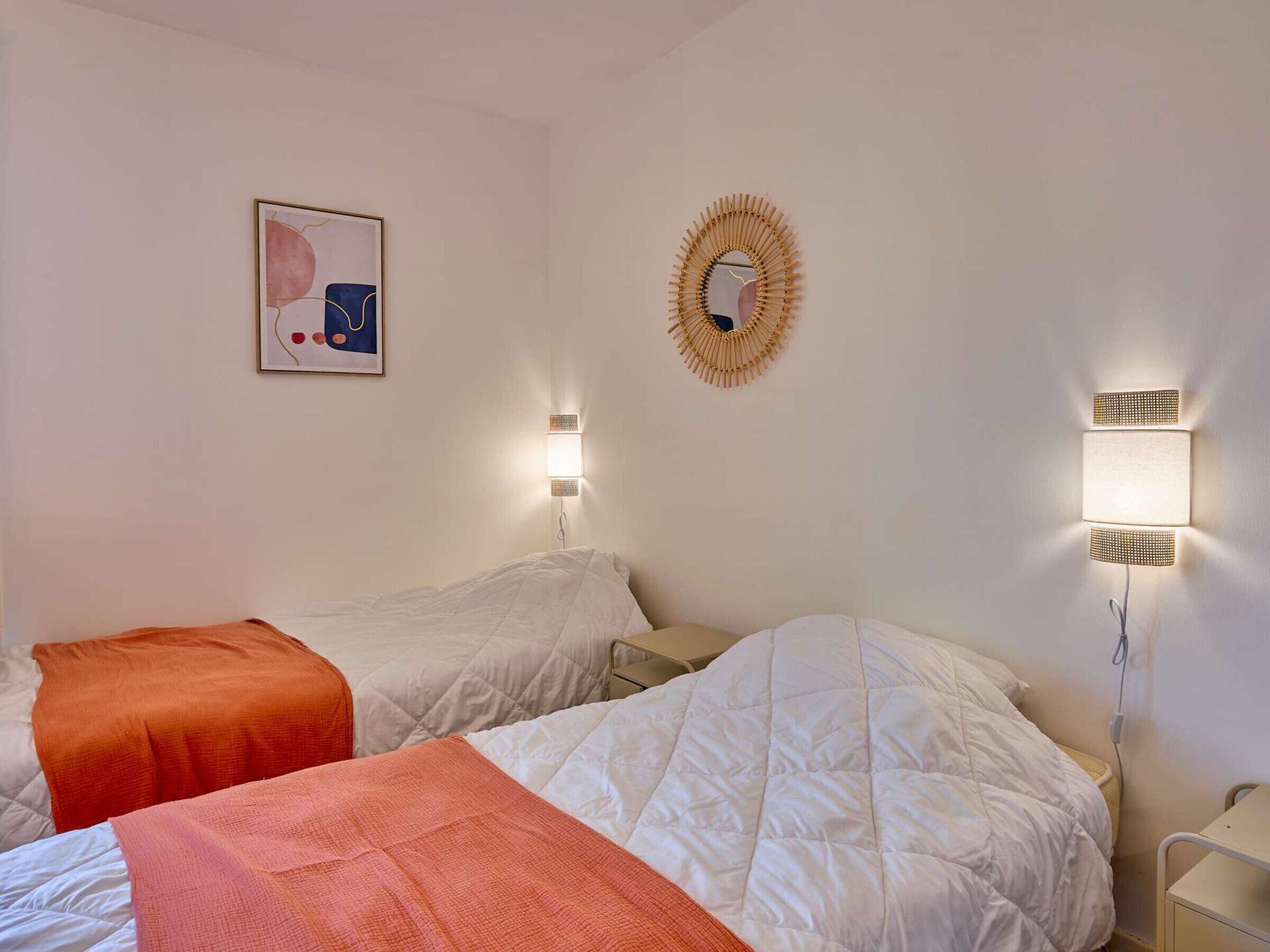 A hotel room with two beds, each with a white duvet and an orange throw blanket. There is a framed abstract artwork on the wall and a decorative mirror above the beds.